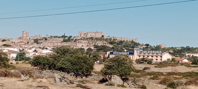 The image shows a panoramic view of the city of Trujillo, Spain, with a castle on a hilltop in the background. The castle is made of stone and appears to be very old. In the foreground, there are some trees and rocks. The sky is clear and blue.

Specifically, the image captures Trujillo from a distance, highlighting its historical architecture and natural surroundings. The prominent castle structure on the elevated terrain suggests a fortified historical settlement. The buildings in the city appear to be constructed with traditional materials, likely stone and tile roofs, indicative of Spanish regional architecture. The presence of a hotel and restaurant named "LAS CIGUENAS" and "RESTAURANTE" indicates a functioning tourist area within or near the historic town. The vegetation, including trees and dry grasses, points to a Mediterranean climate. The overall scene conveys a sense of history and scenic beauty.