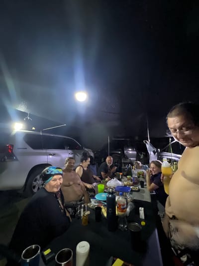 In Karashygan, Kazakhstan, a group of individuals is gathered around a table outdoors at night. The scene is illuminated by a bright overhead light and a smaller light source on a vehicle. People are seated and standing, some interacting with each other while others appear to be eating or drinking.
On the table, there are various items, including bottles of drinks (one appearing to be Fanta), cups, a jar, and plates with food. A blue plastic container is also visible. In the background, several vehicles, including a silver SUV, are parked. The ground is dark, suggesting it's a natural or unpaved area.
The individuals are dressed casually, with some wearing head coverings. The overall atmosphere seems to be a social gathering, possibly a picnic or a casual get-together in an outdoor setting at night.