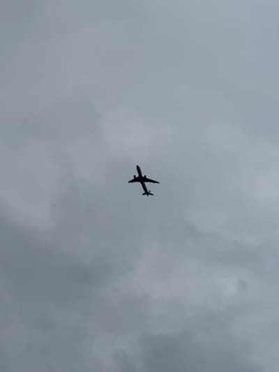 A silhouetted airplane is captured mid-flight against a cloudy, overcast sky. The aircraft is seen from below, with its wings extended and tail visible. The clouds are dense and gray, obscuring any potential landmarks or the ground. The overall impression is one of a routine flight under typical atmospheric conditions. The image does not contain any information that would suggest a location in Rassokha, Russia.
