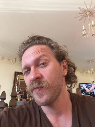 A close-up, rotated shot of a man's face with fair skin, graying curly hair, and a beard. He has an earring in his left ear. The background shows a ceiling with a decorative light fixture, a TV screen showing a person working in what appears to be a kitchen, and a glimpse of a room with a mirror and other objects, suggesting an interior setting. The man is wearing a brown t-shirt. It is not possible to determine the specific location in Тэльмінскі сельскі Савет, Belarus from the given image.