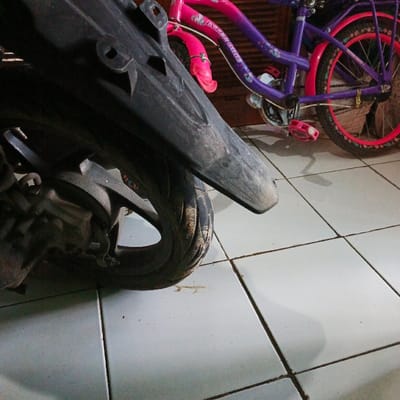 The image shows the rear wheel of a black motorcycle parked on a white tiled floor in Pameungpeuk, Indonesia (latitude: -7.641, longitude: 107.728). The motorcycle's rear fender is visible, extending over the rear tire.  A pink and purple bicycle is parked to the right of the motorcycle, partially obscured by the motorcycle's fender. The bicycle's rear wheel is visible, with its spokes and a red rim. There are no people in the image. The environment is a residential setting, likely a garage or patio.  The image was taken at a low angle, with the camera pointed at the motorcycle's rear wheel. The lighting is dim and diffuse, casting shadows from the motorcycle and bicycle.  The overall impression is one of a casual, everyday scene.