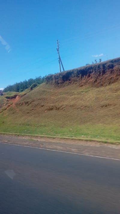A roadside view features a steep grassy hillside against a bright blue sky. A telephone pole stands atop the hill, with wires stretching across the frame. A dark asphalt road occupies the lower portion of the image. The hill exhibits layers of earth and vegetation, with trees clustered along its crest. The overall scene suggests a rural or semi-rural setting captured from a moving vehicle.