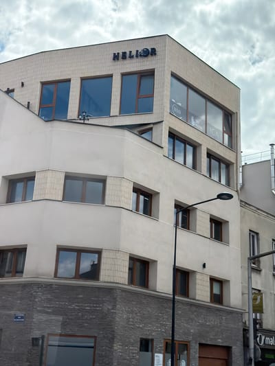 The image shows the exterior of a modern building with a distinctive architectural design. The building features a facade of light-colored, textured material, interspersed with large, dark-framed windows. The windows are of various shapes and sizes, some appearing as long, narrow rectangles, while others are wider and squarish. Some of the windows have interior blinds partially drawn.
The building's design is characterized by its angled sections and the way the windows are recessed or protrude from the main facade. The texture of the building material resembles ribbed paneling on some sections and a smoother finish on others. To the right, a portion of an adjacent building is visible, constructed with a dark, textured material, possibly brick or stone.
There is no activity or event taking place, and no people are visible. The scene appears to be set during the daytime, under a cloudy sky. The clouds are white and fluffy, suggesting fair weather despite being overcast.
Visible text includes "ICA" and "BMI" on signage seen through a large window on the upper left section of the building. Another partial word, "HELIOS," is visible in large, dark lettering on the angled facade to the left. There is also a blue sign with white lettering on the darker building to the right, but the text is illegible. A street lamp with a dark, circular head extends from the building on a thin metal arm.
The overall impression is of an urban setting, likely a business or residential building. The location context provided is Alfortville, France.