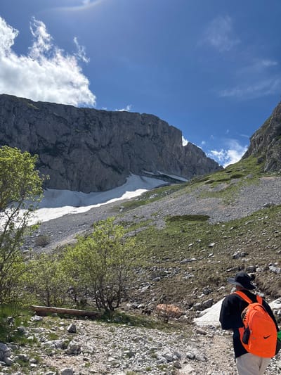 In the mountainous terrain of Pašina voda, Montenegro, a person is walking on a rocky path. The individual is wearing a black hat, a black shirt, light-colored pants, and an orange backpack with a black Nike logo. The environment is characterized by steep, rocky slopes covered with patches of grass and remnants of snow. Lush green trees are scattered across the landscape. In the background, towering grey rock formations dominate the skyline, with a bright blue sky and scattered white clouds overhead.