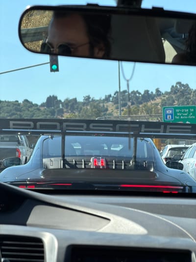 A photograph, taken from the interior of a vehicle, shows a black Porsche with a large rear wing in front of the vehicle. The Porsche's rear lights are visible, as is the word "PORSCHE" on the rear wing. The traffic sign for Tel Aviv-Yafo is visible in the background. In the rearview mirror, a person with dark hair and sunglasses can be seen. The traffic light is green. The location is Jezreel Valley Regional Council, Israel.