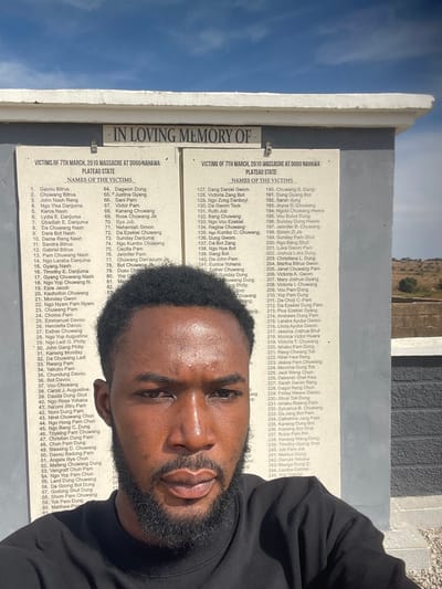 A male subject is depicted in a selfie-style composition, positioned in front of a stone memorial plaque in an outdoor setting, understood to be in Delimi, Nigeria. The subject is a dark-skinned male with a short beard and short, dark, curly hair, wearing a black long-sleeved garment, looking directly at the camera. The memorial plaque, constructed of a light-colored material with dark lettering, is inscribed at the top with "IN LOVING MEMORY OF". Below this, it is divided into two sections, each prominently titled "VICTIMS OF 7TH MARCH, 2010 MASSACRE AT DOGO NAHAWA PLATEAU STATE" and "NAMES OF THE VICTIMS." Numerous numbered names are listed on both sections of the plaque, with some obscured by the subject's head. The plaque is topped by a white concrete overhang. The background features a clear blue sky, and to the right, distant arid landscape with sparse vegetation and uneven terrain is visible. The scene is illuminated by natural daylight.