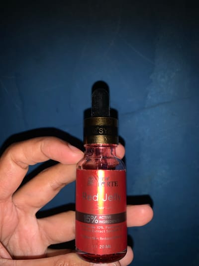 A hand holds a small, red glass bottle with a dropper. The bottle has a gold band around its neck, with some text visible on it. The bottle contains a red liquid, and a label on the bottle reads "NEW FORTE Red Jelly Serum" with "15% ACTIVE INGREDIENTS" below it. The background is a dark blue, textured surface. No specific location in Karangkemiri, Indonesia is identifiable.