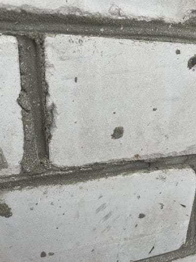 The image shows a close-up view of a weathered white brick wall with dark gray mortar. The bricks have an uneven, slightly rough texture and are stained with scattered dark spots and smudges. The mortar is visibly rough and uneven in places, with some chipped or broken areas revealing aggregate. The lighting suggests an outdoor environment, with subtle shadows indicating the direction of light. The scene is consistent with the exterior of a building, potentially an older structure, in an urban or semi-urban setting. Without further context, a precise location in Kotlas, Russia, cannot be determined from the image alone.