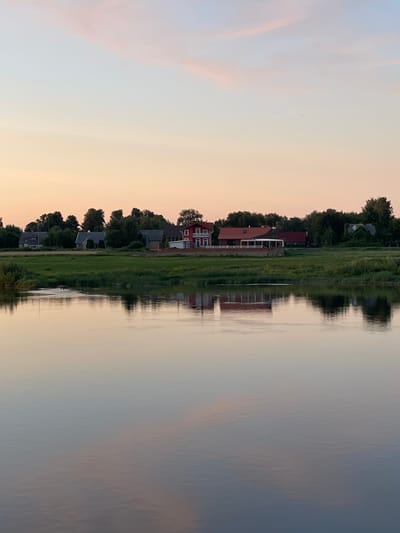 The image depicts a serene river scene at dusk in Sovetsk, Russia. The sky is a soft gradient of pink and pale blue, with wispy clouds. Along the riverbank, a line of houses and trees creates a distinct horizon. The foreground is dominated by the calm surface of the river, which reflects the sky and the structures on the shore with a gentle distortion. The scene is tranquil and devoid of human activity, conveying a sense of peaceful evening in a rural or suburban setting.