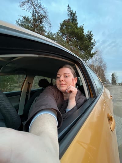 A woman is taking a selfie from the open window of a mustard-yellow car. She is wearing a brown jacket and has her hand touching her face. Outside the car window, trees are visible, and the sky is overcast. A road and some buildings can be seen in the distance, suggesting the location is in or near Voronino, Russia. The interior of the car includes a black seat.