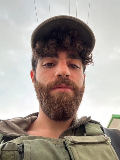 A male individual is depicted in a close-up, low-angle selfie, looking directly into the camera. He has dark, curly hair protruding from under a dark olive-drab cap and a full, reddish-brown beard and mustache. His skin tone is fair to olive. He wears an olive-drab tactical vest with visible pouches and straps, a light brown or grey hooded garment beneath it, and a dark strap of a bag over his left shoulder (viewer's right). The background consists of a predominantly overcast, light grey sky with several thin, dark horizontal lines, likely overhead power or communication cables. A small portion of a structure with green and white elements is visible on the far right. Specific details for geopolitical localization within Brener Regional Council, Israel, are not discernible from the image.