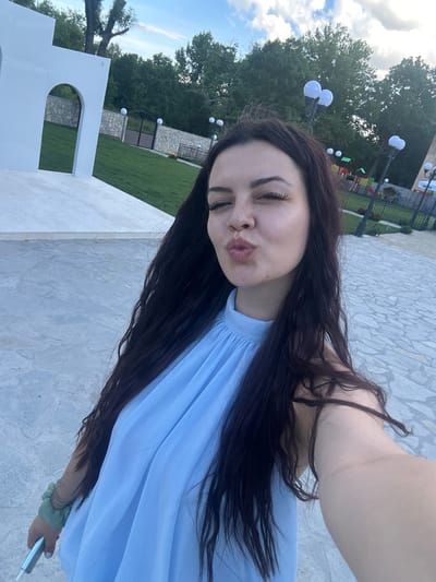 A young woman with long, dark hair and freckles is taking a selfie outdoors. She is wearing a light blue dress and puckering her lips. In the background, there is a grassy area with a stone wall, a gate, and a playground. The location is identified as Zlati Voyvoda, Bulgaria. The overall scene suggests a casual, outdoor setting.