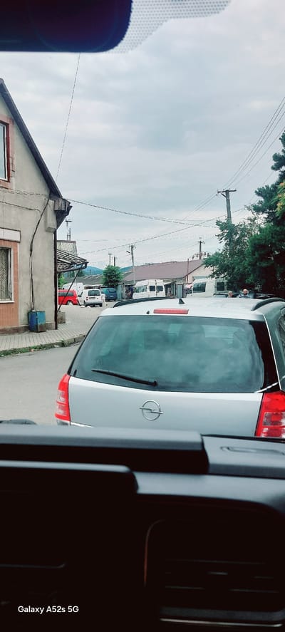 The image shows a street scene in Korolevo, Ukraine, on an overcast day. In the foreground, the back of a silver station wagon is prominently displayed, with its rear window and taillights visible. The car has an Opel logo on its tailgate. Behind the station wagon, a street runs into the distance, lined with buildings and trees. Several other vehicles, including white vans and a red van, are parked or moving on the street. Power lines crisscross the sky, and utility poles are visible. In the background, mountains can be seen in the distance. The overall impression is of a quiet, possibly rural or semi-urban, street with some commercial activity.