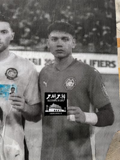 A close-up shot of a monochromatic, halftone-patterned poster, possibly adhered to a wall in George Town, Malaysia. The poster features a man wearing sunglasses and a collared shirt with a possible team logo or emblem on his chest. To the right, a black sticker is applied, displaying the phrase "ALCOHOL IS LIFE" alongside Chinese characters and an architectural graphic. Below the sticker, a blue sticker or graphic is visible. The surrounding environment includes a textured surface, possibly a concrete or weathered wall, adding to the gritty urban feel.