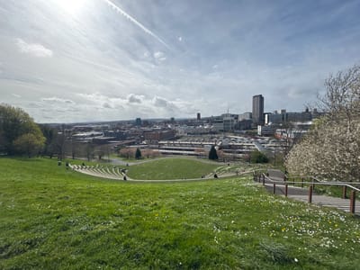 The photograph depicts a panoramic view of Sheffield, United Kingdom, taken from a high vantage point within a park setting, most likely Norfolk Heritage Park. In the foreground, a grassy slope is populated with wildflowers, particularly daisies. A partially visible path with railings leads down the slope on the right. The park features a landscaped amphitheater with seating integrated into the hillside. Scattered individuals can be observed utilizing the park's facilities.

The cityscape in the background includes a mix of low-rise buildings and high-rise towers. The Q-Park Sheffield station and Sheffield Hallam University can be seen in the skyline. Overhead, a bright sun shines through scattered clouds, marked by contrails. The scene suggests a spring day with fair weather.