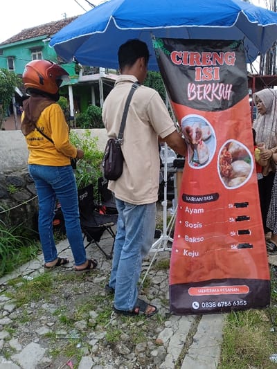 An outdoor scene in Kauman, Indonesia, depicts a street food stall operating under a blue market umbrella. In the center, a person wearing a light brown short-sleeve shirt, blue jeans, and black sandals, with a dark shoulder bag, stands facing away from the camera, positioned behind a vertical red and black banner. The banner advertises "CIRENG ISI BERKAH" (Filled Cireng Berkah), displaying images of cireng with various fillings, a menu listing "Ayam," "Sosis," "Bakso," and "Keju" for 5,000 IDR each, and a contact number for orders. To the left, a person wearing a yellow long-sleeve shirt, blue jeans, and an orange motorcycle helmet stands with their back mostly to the camera, beside a black folding chair. Partially visible on the far right, another person wearing a light-colored hijab appears to be interacting with the stall. The ground is a rough, broken paved surface with patches of grass and weeds. In the background, residential houses with varied colors, including a green one with a brown tiled roof, are visible. A low stone wall is present on the left side of the scene.