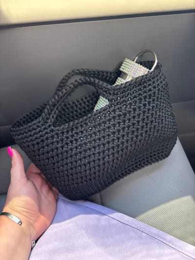 A person's hand, with bright pink fingernails, holds a black crocheted bag. The bag contains what appears to be a silver item encrusted with rhinestones and a silver ring. The background suggests the interior of a vehicle, with a gray leather seat and a lighter gray textured surface above. The person is wearing a light purple garment. There is no indication of the city of Сухарэўскі сельскі Савет, Belarus.