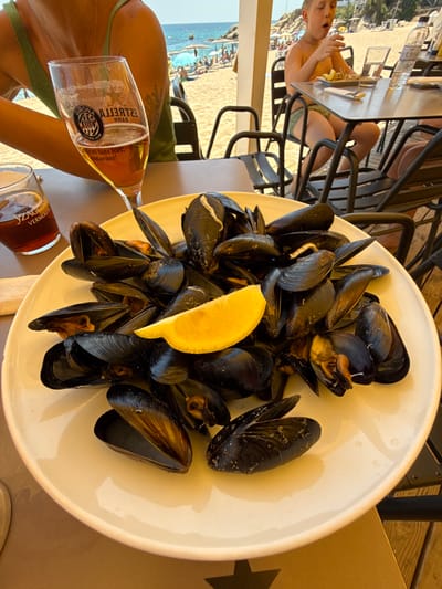 A plate of mussels, a slice of lemon, and a glass of beer sit on a table at a beachfront restaurant in Platja d'Aro, Spain. In the background, a young boy eats at a table, and beyond him, beachgoers relax under umbrellas. The restaurant appears to be an open-air establishment, with wooden beams and chairs visible. The overall scene suggests a casual, seaside dining experience.