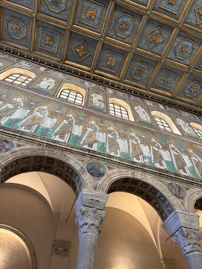 This image showcases the interior of a historical building in Ravenna, Italy. The viewer is looking upwards, capturing the ornate ceiling and walls. The ceiling is designed with repeating geometric patterns of squares and octagons, adorned with gold floral motifs. Adjacent to the ceiling, the wall is decorated with mosaic art, featuring rows of figures with halos above their heads. The windows punctuate the mosaic artwork, providing natural light. A series of arches supported by a fluted column is also visible. The arches are decorated with more mosaic artwork and small portrait medallions.