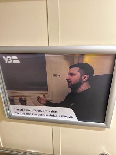 This image displays a vertically oriented, framed poster mounted on a plain, light-colored wall, characteristic of an interior space such as a train carriage. The poster features a photograph of Volodymyr Zelenskyy, President of Ukraine, shown from the chest up in profile, facing left. He has short dark hair and a beard, and wears a dark long-sleeved shirt with an embroidered emblem resembling the Ukrainian trident on the left chest. His right hand is raised in a speaking or gesturing posture. The background of the photograph within the poster includes elements suggesting an interior compartment, such as a window frame and wall details, with the numbers "16 46" visible on a panel. The "УЗ" (Ukrainian Railways) logo is present in the upper left corner of the poster's image. Below the photograph, white text states: "I need ammunition, not a ride. For the ride I've got Ukrainian Railways." The poster itself is enclosed in a silver-colored metallic frame. A metallic fixture is partially visible at the bottom edge of the overall image. The specific location within the city of Kvitneve, Ukraine, cannot be determined from the visual information provided.