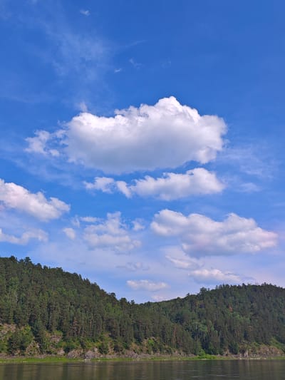 The image depicts a scenic view of the Tayets River in Predivinsk, Russia, under a clear blue sky dotted with fluffy white clouds. A dense, verdant forest covers the steep slopes of the riverbank, with a rocky shoreline visible in places. The water of the river is calm and reflects the sky and surrounding landscape. The scene is natural and undeveloped, conveying a sense of peace and tranquility.
