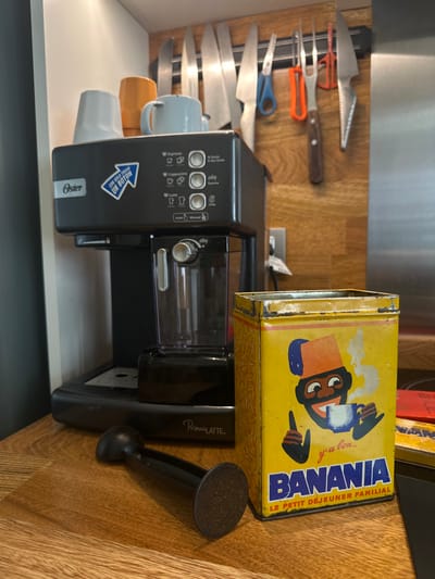 This image depicts a domestic kitchen counter in Acassuso, Argentina. The scene is dominated by a black Oster PrimaLatte espresso machine, positioned centrally on a light-colored wooden countertop. On top of the espresso machine, a light blue mug, an orange mug, and a white or light grey mug are inverted. The machine's control panel displays options for Espresso, Cappuccino, and Latte, with a blue arrow sticker pointing right and stating "CON SÓLO TOCAR UN BOTÓN" (With just one touch of a button). To the right of the machine, a vintage yellow Banania tin, showing signs of wear and rust along its edges, stands upright. The tin features a smiling caricature of a person with dark skin and a red fez, holding a steaming blue cup, with the text "y'a bon.. BANANIA LE PETIT DÉJEUNER FAMILIAL". A black coffee tamper lies horizontally on the wooden countertop in front of the espresso machine. Behind the machine and tin, a magnetic strip mounted on a wooden wall panel holds several knives of varying sizes, a pair of blue-handled scissors, a carving fork with an orange handle, and another red-handled utensil. A white electrical outlet is visible on the wooden wall panel to the right of the espresso machine. No individuals are present, and no active brewing or actions are depicted.