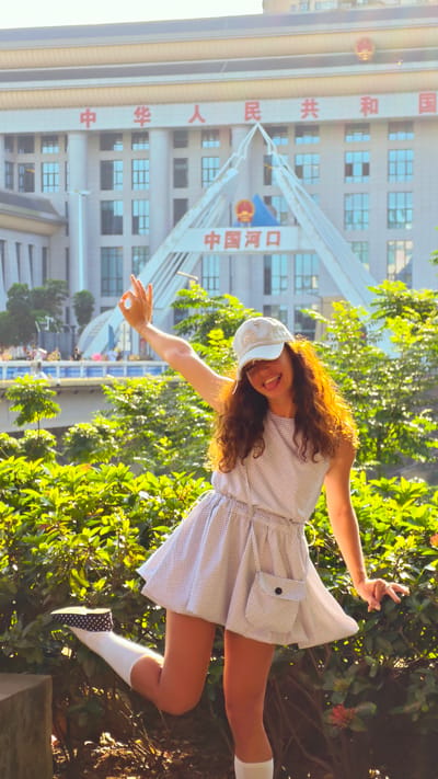 A woman is posing in front of a building in Lao Cai Ward, Vietnam. The woman is wearing a patterned dress, a baseball cap, socks, and shoes. She is standing on one leg and making an "OK" gesture with her hand. The building behind her is white and has Chinese characters on it. There are trees and bushes in the foreground.