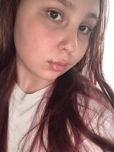 A close-up, diagonal portrait captures the face of a young person, possibly female, with long, dark reddish-brown hair spread around her head. She is lying down, looking directly at the viewer with a neutral expression and dark brown eyes. Notable features include a silver barbell piercing above her left eyebrow and a silver hoop septum piercing on her nose. Her lips are full and slightly parted. Her skin is fair, showing some natural texture. A simple white t-shirt or fabric is visible on her shoulder and neck area. The background consists mainly of her hair and indistinct light-colored bedding or fabric, suggesting an indoor setting. The lighting is soft and even. This image is provided with the location context of Lyman, Ukraine.