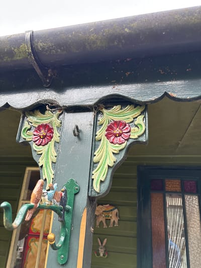 This image captures a forensic view of an exterior building structure in Stiperstones, United Kingdom, specifically focusing on a section of a dark green facade. A dark, moss-covered gutter system with a visible support bracket spans the top. Below the gutter, a dark green scalloped eave overhangs a prominent dark green vertical wooden beam. Attached to this beam are two ornate carved wooden corbels, each depicting a red flower with a yellow center encircled by light green leaves, set against a dark green background. Cobwebs are evident around these decorative elements and under the eave. A small, dark metal ring is secured to the beam between the corbels. Further down the beam, a dark green metal coat hanger with three hooks is mounted. Perched on the upper hook are three small, carved wooden birds: one brown, one blue, and one black, positioned in close proximity. To the right of the beam, on the dark green wall paneling, two wooden decorative items are affixed: an intricately carved elephant with colorful detailing above, and a smaller carved rabbit head with red floral accents below. A window with a dark frame is partially visible on the far right, featuring multi-colored, likely stained, glass panes (orange, amber, clear) in its upper section, and a reflective lower pane showing a distorted reflection of a light blue garment. The overall structure displays weathering, including moss, lichen, and rust-colored marks. No individuals are directly present.