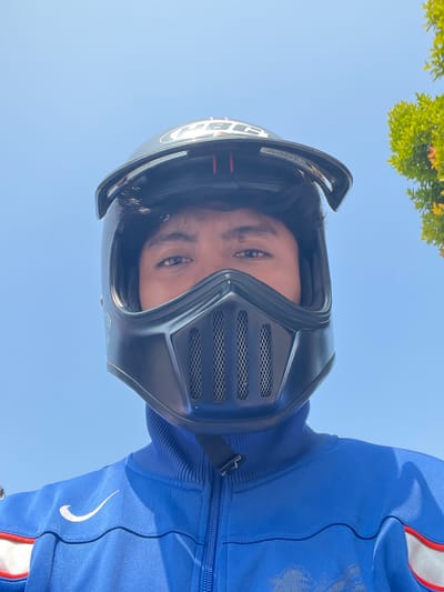 A person wearing a black full-face helmet and a blue jacket with a Nike logo is pictured from a low angle against a clear blue sky, with a portion of a green tree visible in the upper right corner. The individual's eyes are visible through the helmet's visor, and the helmet has a distinct mask with a mesh grille. The jacket appears to be a tracksuit jacket with red and white accents on the sleeves. The image was captured in Batang, Indonesia.