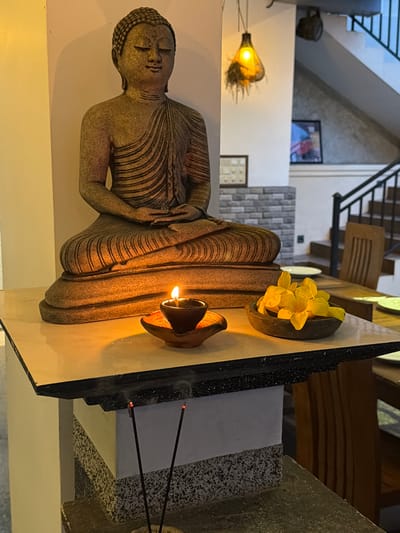 This image features a statue of a reclining Buddha, a common religious and artistic motif. The statue is made of a textured, stone-like material and is adorned with carved lines that suggest drapery. Its serene expression and peaceful posture evoke a sense of tranquility.

Adjacent to the Buddha statue, a small oil lamp burns, casting a warm, flickering glow that illuminates the sculpture and the immediate surroundings. The flame is small and contained within a dark, cup-shaped holder.

Further to the right of the Buddha, three incense sticks are burning, emitting wisps of smoke that curl upwards. The glowing red tips of the incense sticks indicate they are actively burning.

In the background, there are hints of an interior setting. A staircase with a dark railing is visible, along with wooden elements and brickwork, suggesting an indoor space like a temple, monastery, or a home decorated with religious artifacts. The lighting in the scene is predominantly warm and ambient, likely from the oil lamp and the incense, creating a meditative atmosphere.

The overall mood is one of peace, reverence, and spiritual contemplation. The presence of the Buddha statue, the lamp, and the incense strongly suggests a religious or meditative context. The image appears to be taken indoors, likely during the evening or night, given the emphasis on artificial light sources. The location context of Rumassala, Sri Lanka, is significant as Sri Lanka is a predominantly Buddhist country with a rich tradition of Buddhist art and worship.