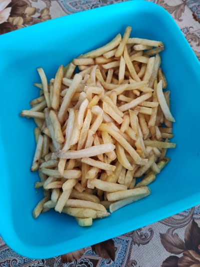 A blue plastic container is filled with a pile of pale, slightly golden French fries. The container is sitting on a surface covered with a patterned tablecloth, featuring floral and decorative elements in shades of brown and beige. The photo appears to be taken indoors. There is no evidence to suggest the location of Musachevo, Bulgaria.