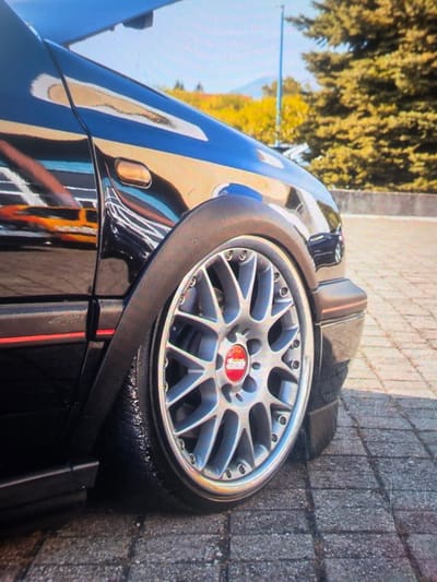 This is a close-up, eye-level, outdoor, daytime, medium shot of a black car's front fender and wheel. The car is parked on a brick-patterned surface. The car's body is highly reflective, showing a distorted reflection of an orange vehicle and other elements. The wheel is silver with a red and white BBS logo in the center. The tire appears wet. In the background, there are trees and a blue sky. The scene is located in Maasvlakte, Rotterdam, Netherlands.