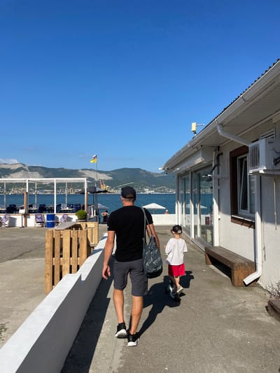 The image depicts two individuals standing outdoors on a sunny day, possibly in a coastal or lakeside area, given the presence of water and mountains in the background. The sky is a clear, bright blue, suggesting fair weather and likely daytime.

In the foreground, a person wearing a black t-shirt, grey shorts, a black cap, and black and white shoes is facing away from the camera. They are carrying a patterned tote bag over their left shoulder. To their right, a younger person is visible, wearing a white t-shirt and red shorts, also with a cap. This individual appears to be walking or standing near a wooden bench attached to the side of a building.

The building features white siding, a white gutter system, an air conditioning unit, and windows. One window reflects the surrounding environment. The ground is paved with concrete. In the mid-ground, there is a railing and beyond that, a body of water with mountains in the distance. Several chairs and blue barrels are arranged in an outdoor seating area. A flag pole with a flag is also visible.

Wooden pallets are stacked near the foreground. The overall atmosphere suggests a relaxed, outdoor setting, possibly a cafe or public space near a waterfront. The lighting indicates it is likely late morning or early afternoon. There is no visible text in the image.