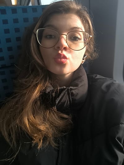 The image displays a close-up, waist-up portrait of a young adult female. She has long, wavy, light brown hair flowing over her left shoulder and chest. Her face exhibits fair skin, gold-rimmed cat-eye glasses, and red lipstick on her pursed lips. A dark earbud is visible in her right ear. She is dressed in a black puffer jacket. The background to her left features a blue seat with a repeating square grid pattern, while the right side shows a neutral-toned interior surface, possibly a wall or window reflecting light. The lighting suggests a natural light source from the right side of the frame. The scene is an interior location, identified as Kopanitsa, Bulgaria.