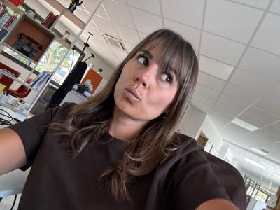 A woman with long brown hair and bangs is taking a selfie. She is wearing a brown sweater. The background is a bright indoor space with white tiled floors and a bookcase filled with books and decorative objects. The scene is not explicitly identifiable as Les Arcs, France based on the provided image alone.