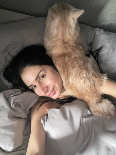 The image displays a close-up view of an individual and a domestic cat in a bed, located in Syasstroy, Russia. A person with dark hair is lying on their back, their head positioned in the lower-left portion of the frame. Their face is turned towards the camera, showing a faint smile. An orange tabby cat is resting on the person's upper torso and neck area, oriented with its back towards the viewer. The cat's head is turned away from the camera, facing the upper-left. Both the person and the cat are partially covered by light gray bedding, which also forms the immediate background.