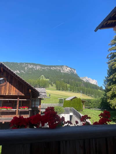 The image depicts a sunlit mountain landscape in La Ila - Stern - La Villa, Italy. In the foreground, a wooden balcony railing is visible, with vibrant red flowers in bloom above it. To the left, a rustic wooden house with a circular window and flower boxes adorns the scene. Behind the house, a lush green meadow stretches towards a wooded mountain range. Ski lifts are visible on the mountainside, suggesting a recreational area. Tall evergreen trees frame the right side of the image, and the sky is a clear, bright blue with a faint contrail. The overall impression is one of a serene alpine setting on a clear day.