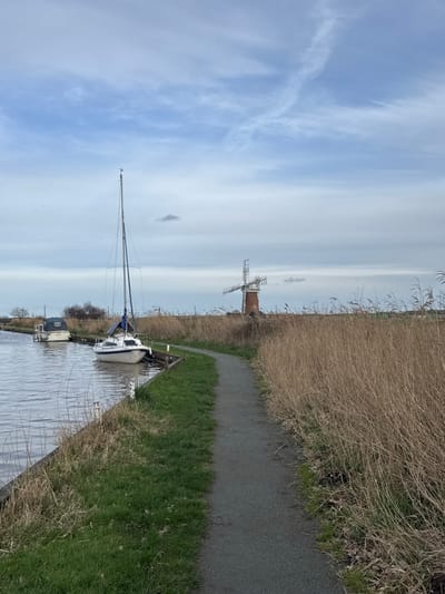 The image displays a rural waterside scene in North Norfolk, United Kingdom, specifically near St Benet's Abbey. A dark grey, unpaved path curves from the foreground towards the middle ground, running parallel to a calm waterway on its left. The path is bordered by short green grass on the left and a dense field of tall, dry, light brown reeds on the right, which extends to the horizon.

Moored along the waterway on the left is a white-hulled sailboat with a tall, slender mast, a blue tarp covering part of its boom, and a visible solar panel on its deck. A small white bird is perched on the masthead. Further along the waterway, partially obscured, a smaller light-colored motor cruiser is also moored. In the background, to the right of the center and rising above the reeds, stands a red brick tower windmill with a white cap and a multi-bladed fantail. The sky is overcast with scattered white and light grey clouds, including a faint contrail. No human figures are present.