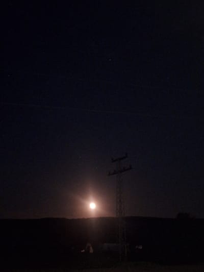 The image shows an electrical tower silhouetted against the night sky in Dolni Chiflik, Bulgaria. A bright light source is visible to the right of the tower, possibly a streetlight or the moon. The surrounding environment appears dark with a vertical dark mass of trees or buildings on the right of the tower/light.