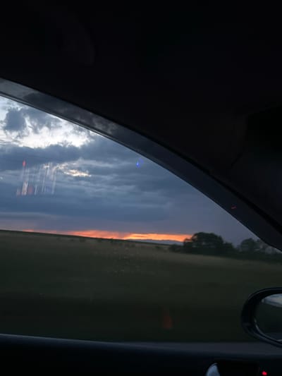 The image presents a view from inside a vehicle, looking out of a side window at either a sunset or sunrise. The dark interior of the car, including parts of the door frame and possibly the roof lining, forms the top and bottom borders of the frame, with a curved edge delineating the window.
Outside, the sky is a dramatic focal point. The upper portion of the visible sky features heavy, dark clouds with patches of lighter, whiter clouds interspersed. Towards the horizon, a brilliant, intense band of orange and red light stretches across, indicating the position of the sun. Below this vivid sky, a dark, rural landscape of fields and silhouetted trees is visible.
Reflections on the car window add complexity to the scene. Prominently in the lower part of the window, a light-colored reflection appears to be signage or a structure, possibly with vertical elements and letters that resemble "ESSO" or a similar commercial brand, hinting at a nearby petrol station or business. Other faint blue and red light reflections are scattered across the window, likely from internal vehicle lights or external sources. On the far right, reflections of a side mirror and an interior vehicle component with a small red indicator light are discernible.
The image captures a moment of travel during dusk or dawn, showcasing the striking natural beauty of the sky over a rural setting, augmented by reflections of the immediate surroundings and vehicle interior. The location context is Ботево, Bulgaria.