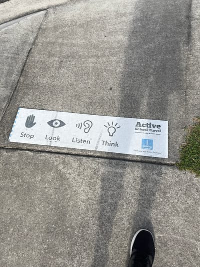 This image captures a concrete sidewalk or path in Riverhills, Australia, under bright daytime conditions. A long, white, rectangular sign or decal is placed on the ground, positioned vertically along the edge where the concrete meets a small patch of green grass.

The sign is an "Active School Travel" initiative, likely promoting road safety for pedestrians. It features a sequence of icons and accompanying text, reading from bottom to top: a hand icon with the word "Stop," an eye icon with "Look," an ear icon with "Listen," and a lightbulb icon with "Think."

At the top right section of the sign, the prominent text "Active School Travel" is displayed. Below this, smaller text reads "Be active. Be safe. Be independent." A blue logo, identified as the "Brisbane City" logo, is present, with the phrase "Dedicated to a better Brisbane" printed beneath it, reinforcing the local context.

A person's foot, wearing a black athletic shoe with a white sole, is partially visible in the upper right corner of the frame, indicating a pedestrian is standing close to the sign. The concrete ground shows typical wear and shadows, consistent with an outdoor urban or suburban environment.