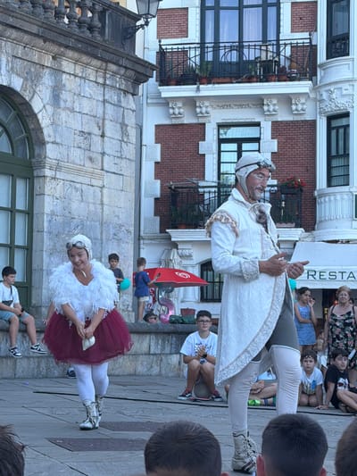 The image depicts an outdoor performance on a paved plaza, likely in Lekeitio, Spain, given the architectural style of the surrounding buildings which are characteristic of coastal Basque Country. In the foreground, two performers are center stage. The performer on the left is dressed in a fluffy white top with a feathered trim and a dark red, full skirt, paired with white stockings and ornate boots. She is wearing goggles and has a playful expression. To her right, a male performer stands in an elaborate white and cream costume featuring intricate embroidery and fringed epaulets. He wears a cap with goggles and appears to be speaking, gesturing with his hands. Behind them, children and adults are seated, some watching the performance, others engaged in their own activities. A red umbrella is visible in the middle ground, and a building with a "RESTA" sign suggests commercial establishments are nearby. The architecture features stone facades and balconies, typical of older European towns. The lighting indicates it is daytime.