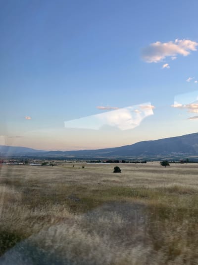 The image depicts a landscape with a combination of natural and built environments. In the foreground, there is what appears to be a field of dry grass or hay. Scattered throughout the field are a few trees. Adjacent to the field is a line of buildings that suggest the edge of a town or city. Beyond the buildings, there is a mountain range in the background. The sky above is blue with some clouds, and there are reflections visible, possibly from a window or other reflective surface through which the scene is viewed. This scene is located in the city of Prosotsani Municipal Unit, Greece.
