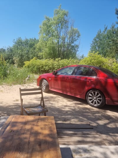 A red four-door sedan is parked on a dirt ground surrounded by dense green foliage and trees under a clear blue sky. In the foreground, a wooden table and chair are set up, suggesting an outdoor seating area. The car's right side is visible, with its wheels on the ground. A small metallic object, possibly a piece of plumbing or a marker, is partially visible near the edge of the vegetation. The ground is sandy and covered with scattered leaves and shadows from the trees. The scene appears to be in a rural or semi-rural area.