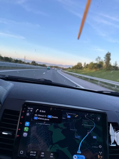 The image shows a first-person perspective from inside a car driving on a highway. The dashboard is visible in the foreground, featuring a large infotainment screen displaying a navigation map. The map shows a route leading to "Malo Buchino" and highlights streets like "ул. Малобучински път". The time is displayed as 20:44, and the speed is 88 km/h. Outside the car, a highway stretches into the distance, with trees and hills visible on the sides. The sky is clear and blue, indicating it is late afternoon or early evening. The overall scene depicts a journey on a highway in Bulgaria.