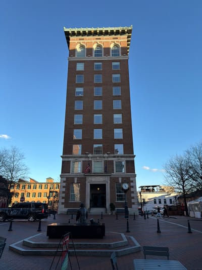A tall, narrow, brick building with several rows of windows stands prominently against a clear blue sky. The building's design includes a decorative top with archways and detailed edging. A clock is visible on the side of the building near the entrance. Below the building, there's a brick-paved area with statues, benches, and street lamps. Other buildings can be seen in the background.
