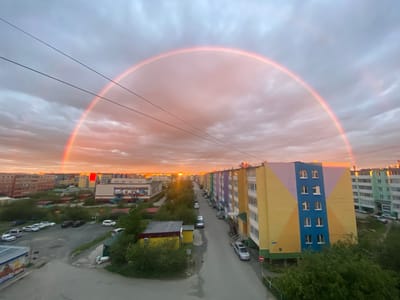 This is a high-angle, wide-shot, eye-level photograph of a residential district in the city of Udachny, Russia, during what appears to be a sunset. A prominent rainbow arcs across the sky, its colors softened by the overcast, pinkish-orange clouds. Two parallel power lines stretch across the upper left portion of the frame, bisecting the rainbow.
The urban environment consists of multi-story apartment buildings with colorful facades, particularly a prominent yellow, blue, and pink building on the right, and a green building behind it. Many windows are visible on the buildings, some open. A street runs through the scene, lined with parked cars, including several white sedans, a silver minivan, and a dark SUV. Green trees and bushes are present along the street and surrounding the buildings, suggesting a temperate or boreal climate. In the foreground, a grey asphalt lot is visible, also with parked cars. A small, single-story yellow building with a dark roof is situated among the trees to the left. The overall impression is of a typical, albeit colorful, urban landscape under a dramatic sky.