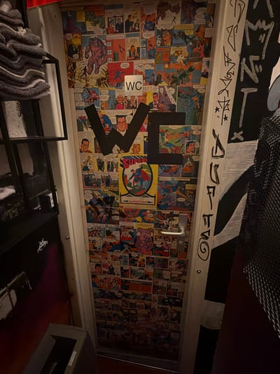 The image shows a wall covered with comic book pages, likely Superman comics, in what appears to be a bathroom in Ulm, Germany. A black 'W' is painted on the wall, partially obscuring the comics. There is a metal WC sign. The wall is located above a white-painted fixture. Below the fixture is a towel rack with towels and a dark-colored shelf. Above the comic strip wall, there is graffiti. A "NO TRASH" sign is on the garbage receptacle to the right of the shelf.