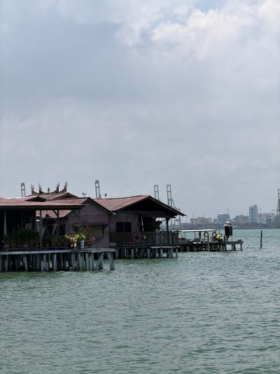 The image shows a stilt house at Chew Jetty, George Town, Malaysia. The house is constructed over the water, supported by wooden piles. It has a corrugated iron roof and walls, with decorative elements on the roof's edges. Plants and flowers adorn the house's platform. In the background, there are modern buildings and harbor cranes. A person is seated on a platform nearby, visible in the distance. The sky is overcast, and the water is calm.